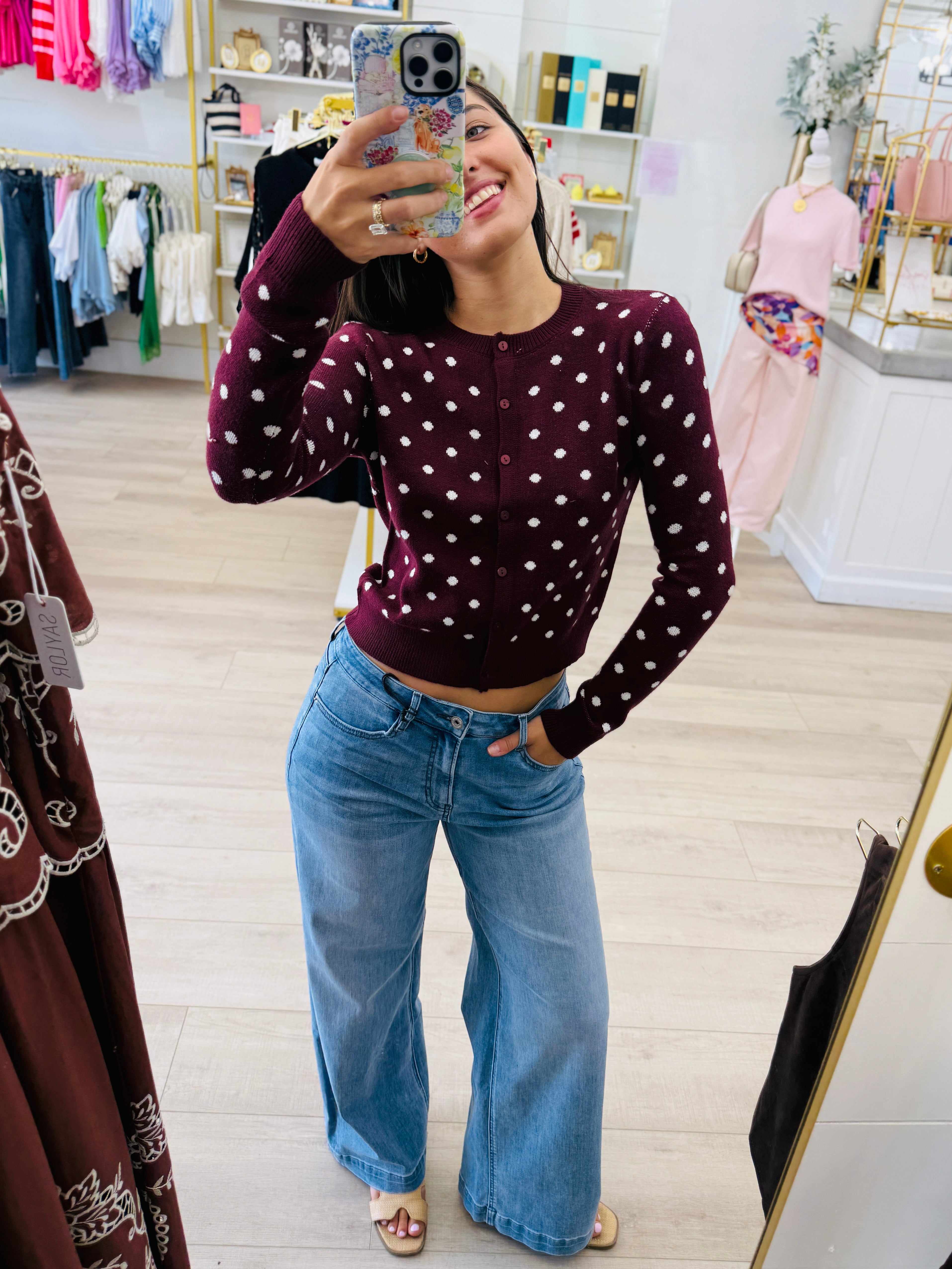 A woman takes a mirror selfie in a clothing boutique, wearing a dark burgundy long-sleeve cardigan with white polka dots and light-wash blue wide-leg jeans.