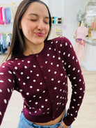 A woman wearing a burgundy cardigan with white polka dots and buttons, paired with blue jeans, in a brightly lit boutique setting.
