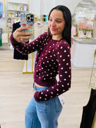 A woman taking a mirror selfie in a boutique, wearing a burgundy long-sleeve crop top with white polka dots and light blue jeans.