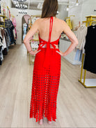 A woman stands with her back to the camera wearing a bright red open-knit maxi dress in a clothing boutique. The dress features a halter-style back with cutouts and a long, fringed lower skirt.