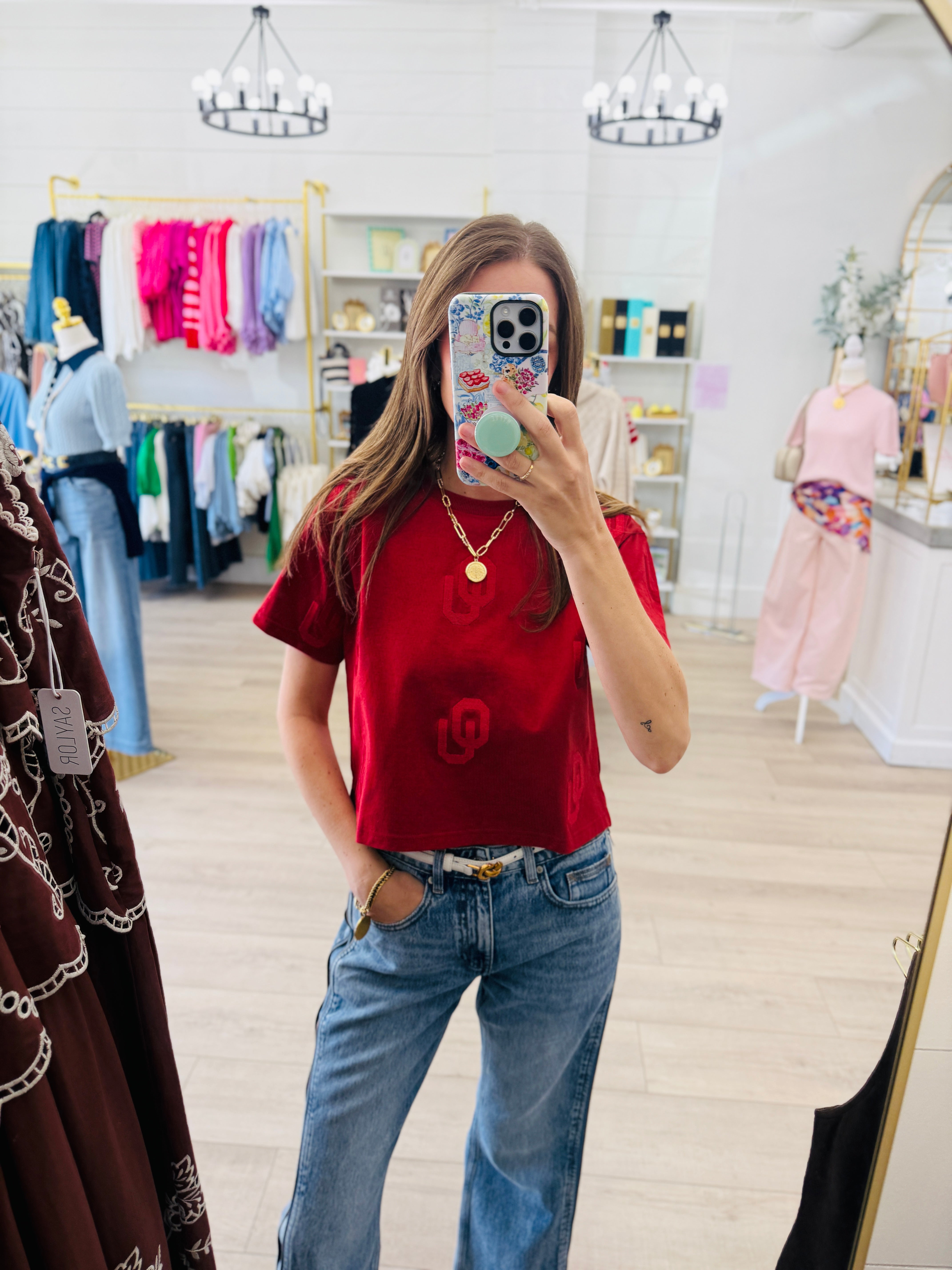 A woman takes a mirror selfie in a boutique, wearing a red short-sleeve t-shirt with a tonal circular logo and blue jeans.