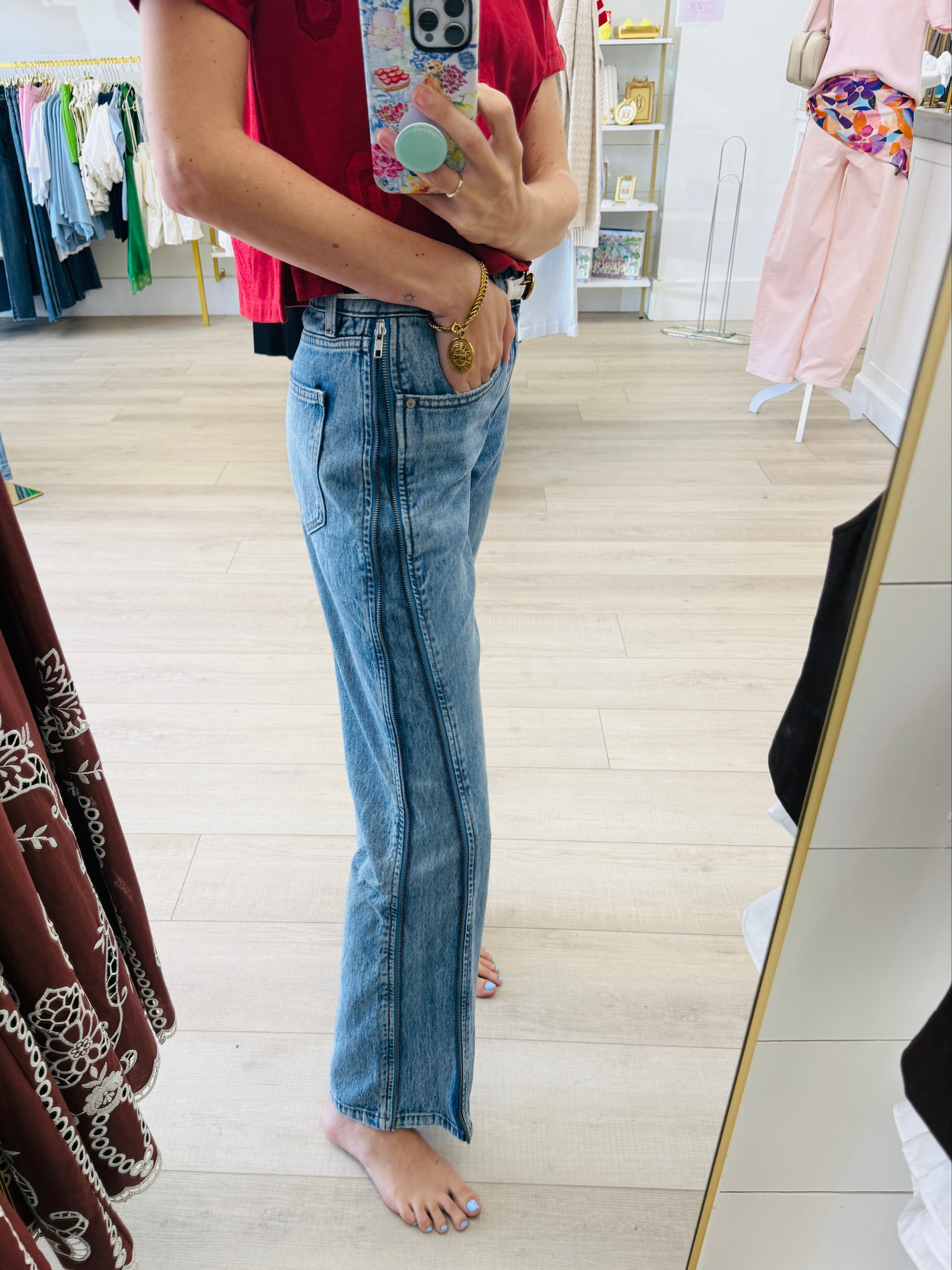 A person taking a mirror selfie in a clothing boutique, wearing light-wash blue flare jeans with a unique vertical seam detail down the front, paired with a red t-shirt.