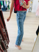 A person taking a mirror selfie in a clothing boutique, wearing light-wash blue straight-leg jeans and a red t-shirt. The boutique has light wood floors, a clothing rack in the background, and a large mirror with a gold frame.