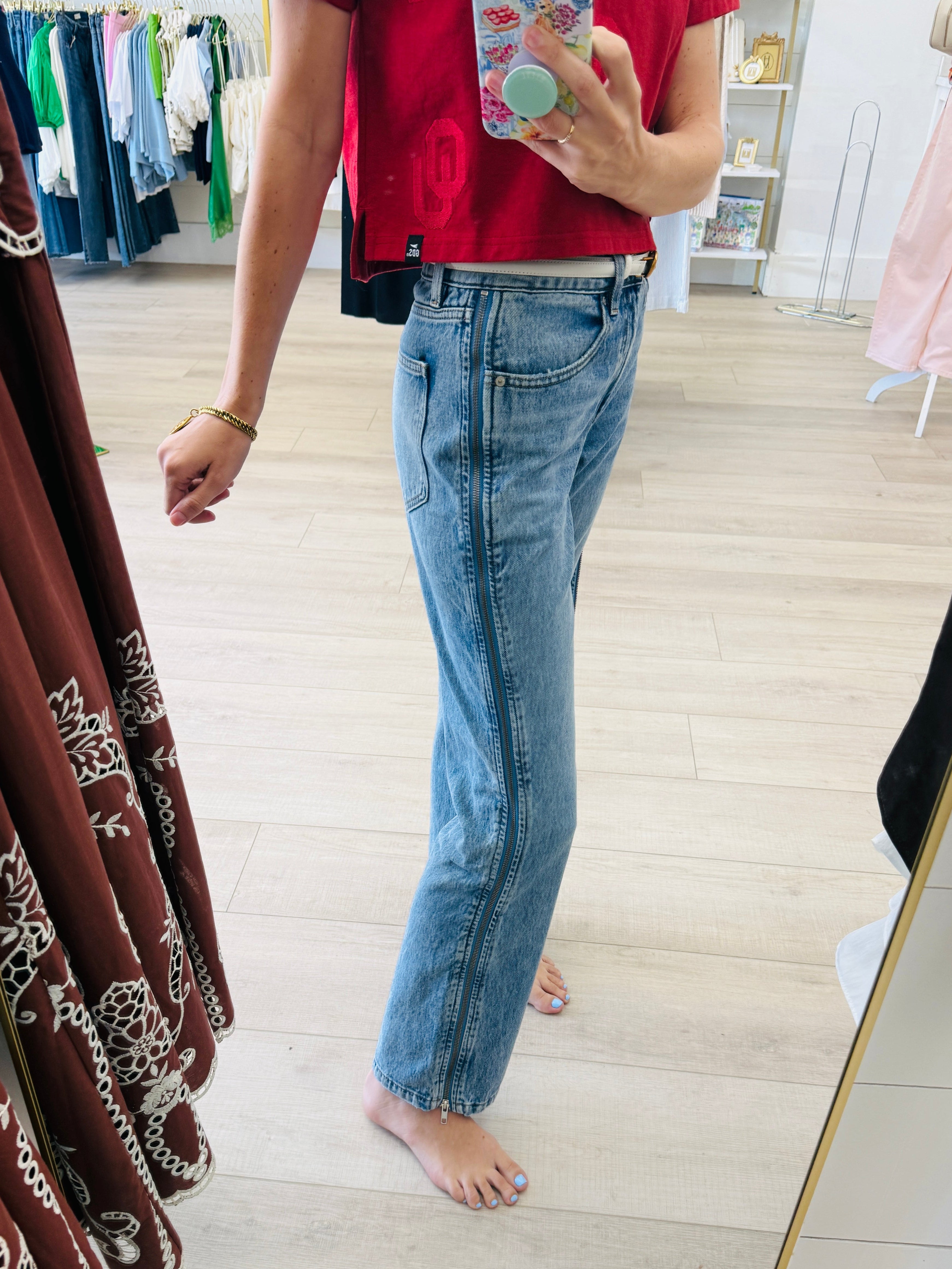 A person taking a mirror selfie in a clothing boutique, wearing light-wash blue straight-leg jeans and a red t-shirt. The boutique has light wood floors, a clothing rack in the background, and a large mirror with a gold frame.