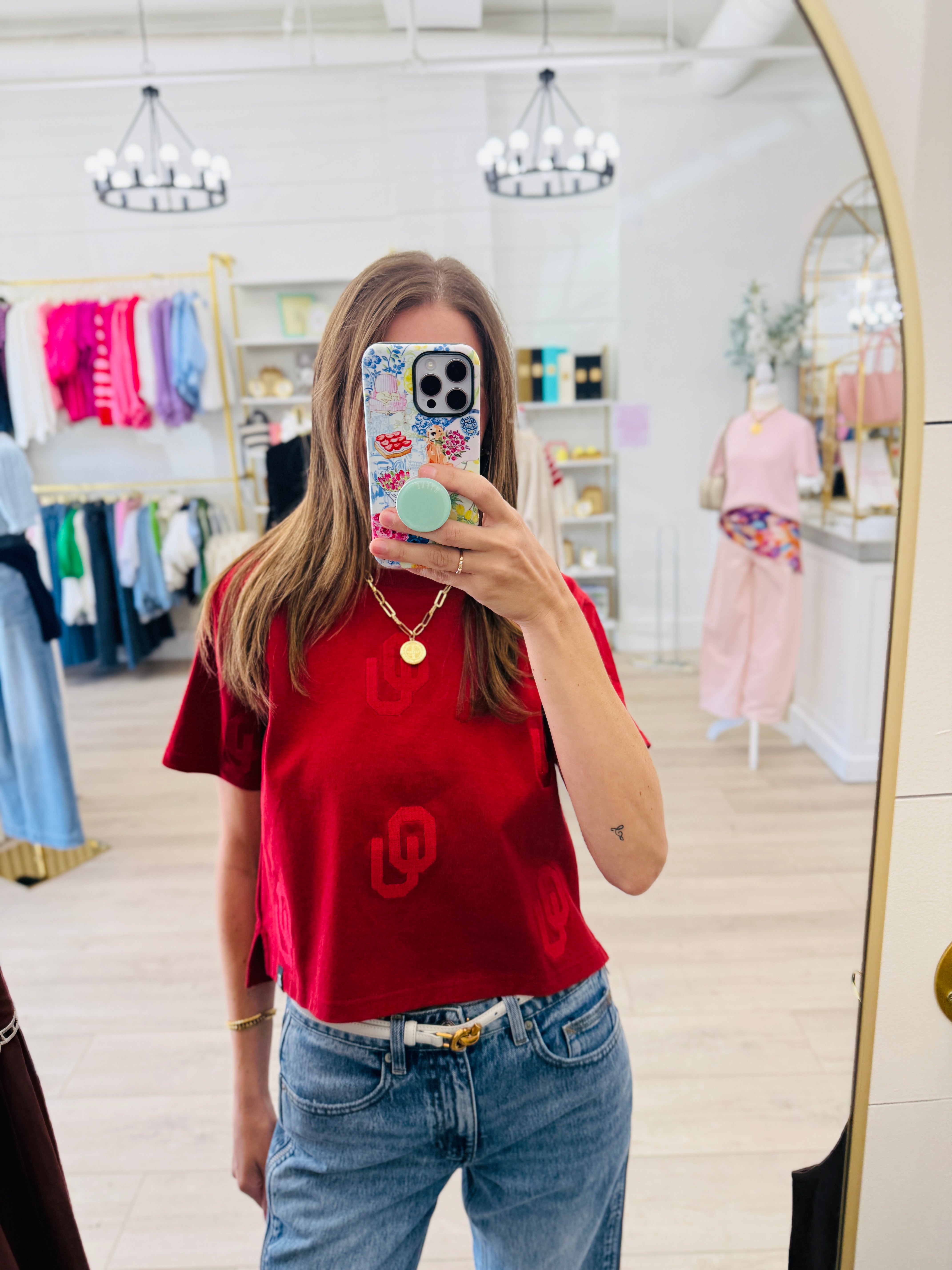 A woman takes a mirror selfie in a boutique wearing a bright red short-sleeve t-shirt with a large red 'U' logo on the front, paired with blue jeans.