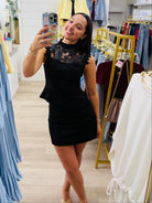 A woman takes a mirror selfie in a clothing boutique wearing a black sleeveless mini dress with lace detailing on the bodice. The boutique features a large gold-framed arch mirror, light wood floors, and racks of clothing in blue, yellow, and white.