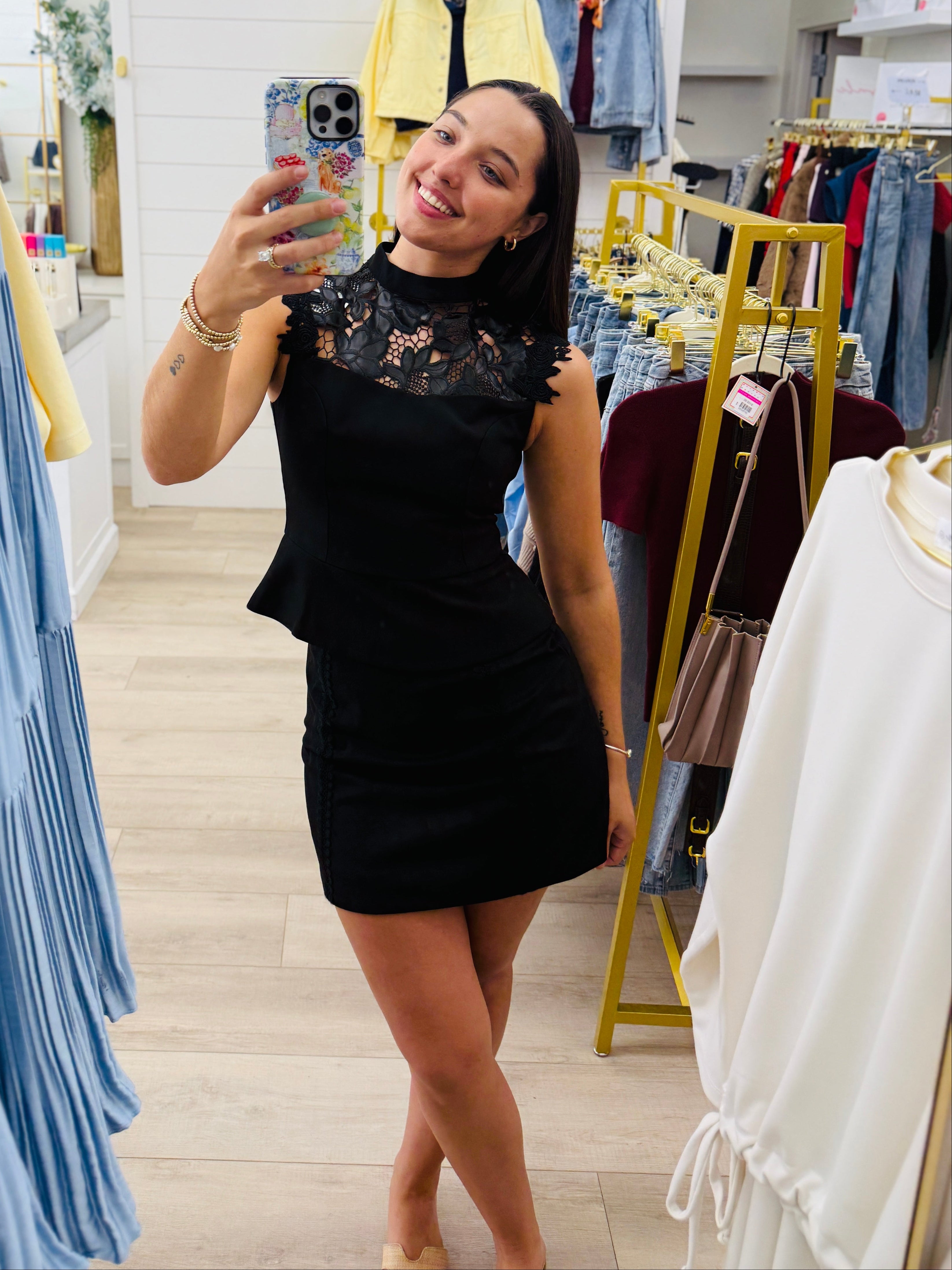 A woman takes a mirror selfie in a clothing boutique wearing a black sleeveless mini dress with lace detailing on the bodice. The boutique features a large gold-framed arch mirror, light wood floors, and racks of clothing in blue, yellow, and white.
