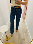 A person taking a mirror selfie in a hallway wearing dark blue straight-leg jeans, a tan belt with a gold buckle, and a light beige top.