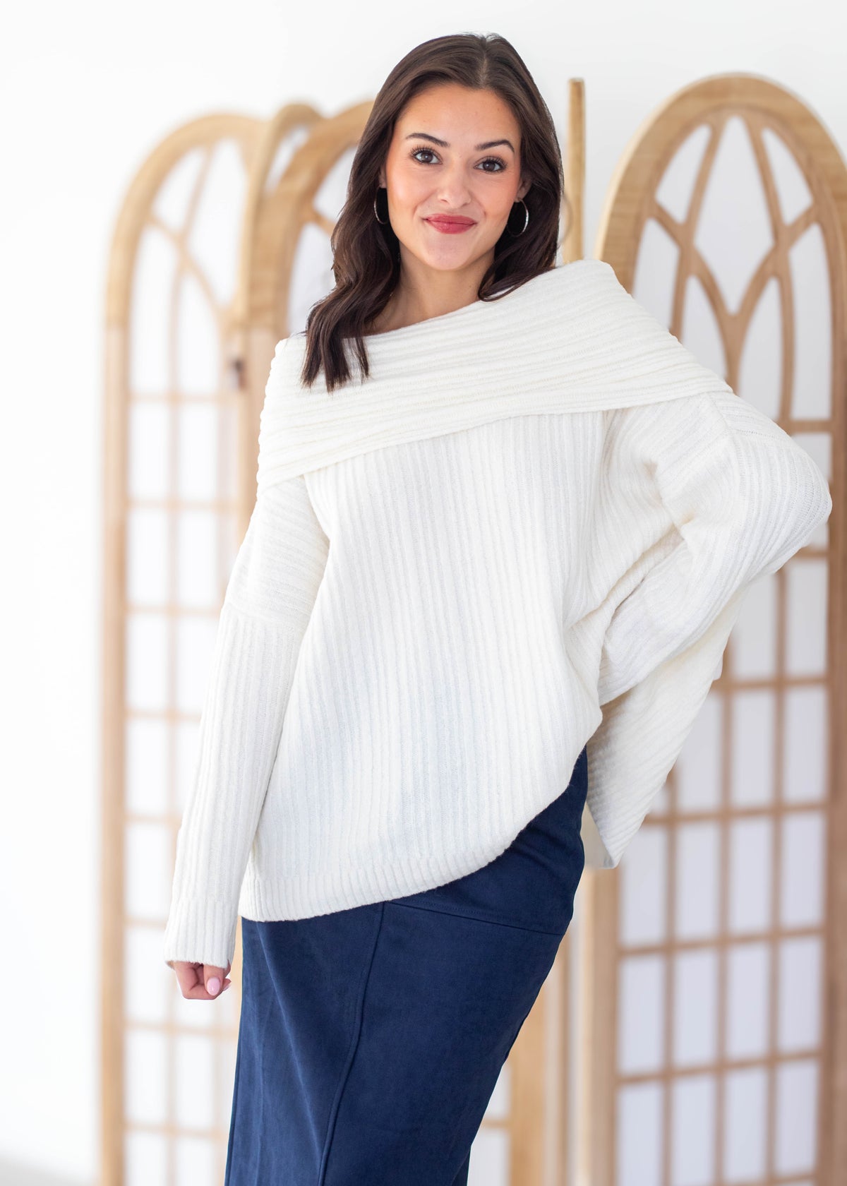 A woman wearing a white ribbed knit sweater with an asymmetrical draped neckline and navy blue skirt, standing in front of a light wood folding screen.