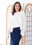 A woman modeling a ribbed off-the-shoulder white sweater paired with a navy blue skirt, standing in front of a light wood folding screen.