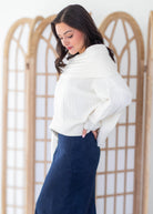 A woman wearing a ribbed white knit wrap sweater and a navy blue skirt, standing in front of a light wood folding screen.