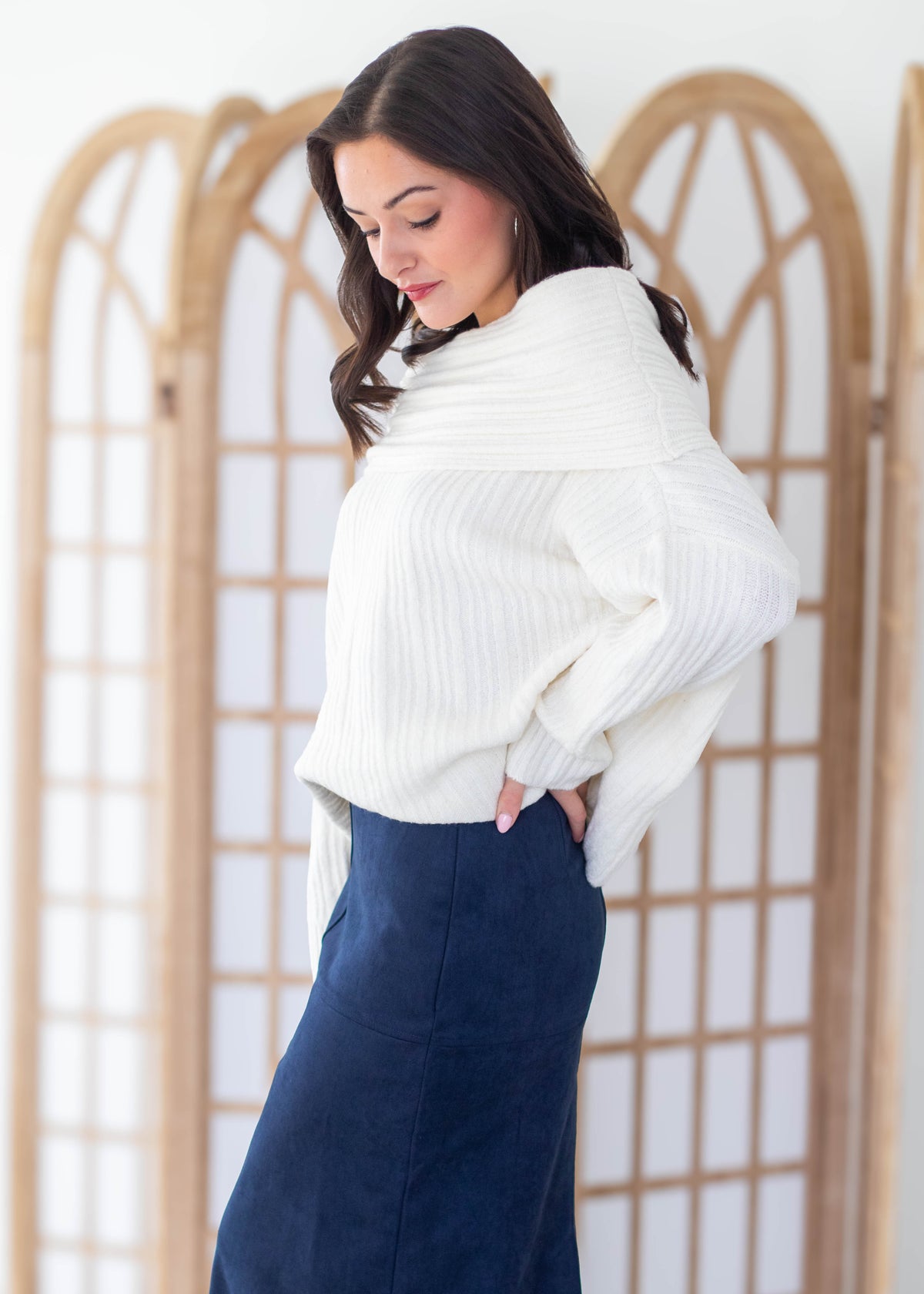 A woman wearing a ribbed white knit wrap sweater and a navy blue skirt, standing in front of a light wood folding screen.