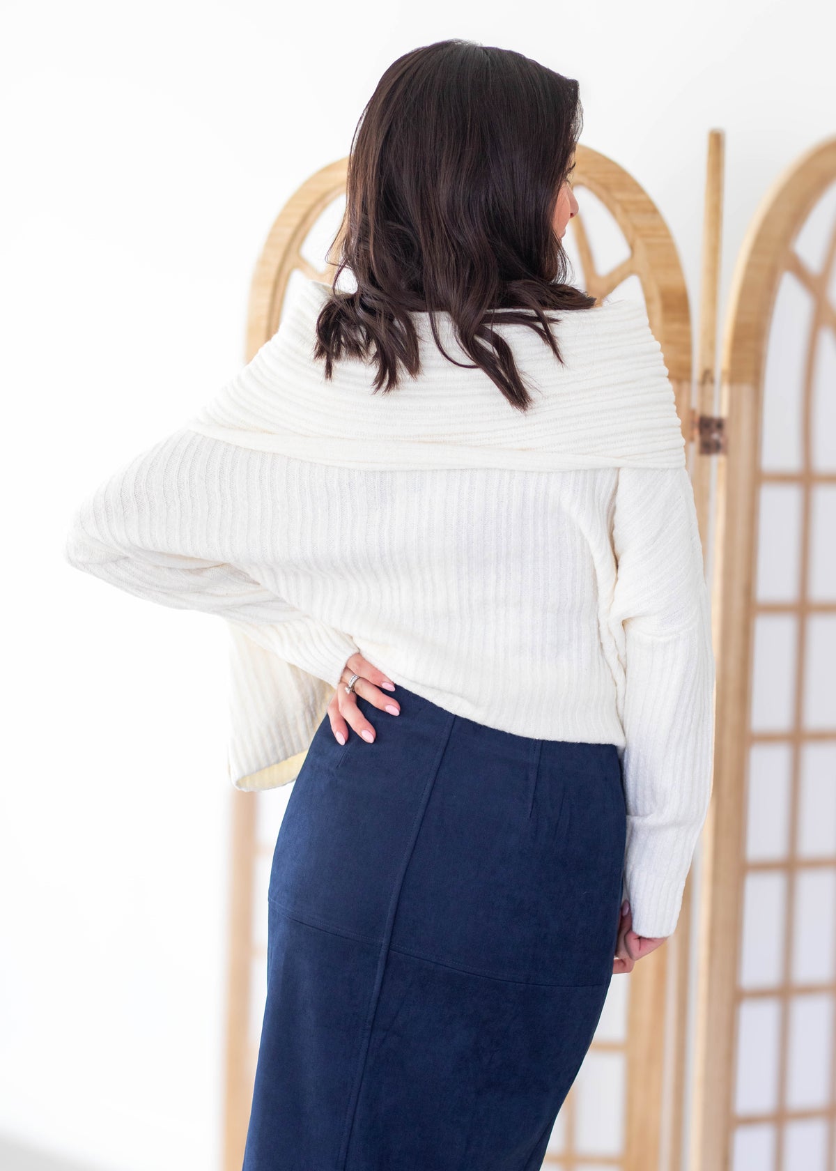 A woman shown from the back wearing a cream ribbed knit wrap sweater and a navy blue skirt, standing in front of a light wood folding screen.