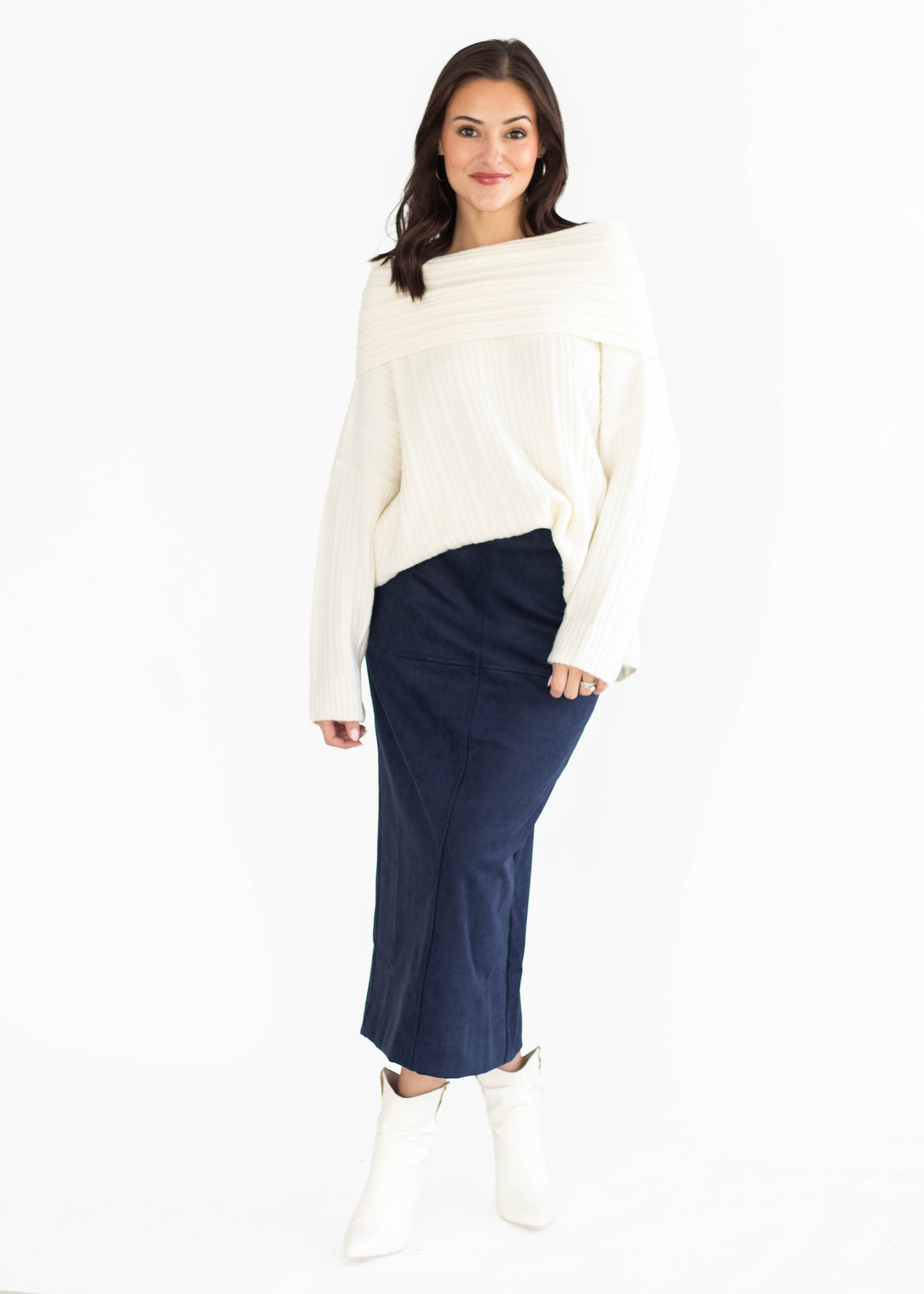 A woman modeling a cream off-the-shoulder knit sweater and a dark navy midi skirt, paired with white ankle boots, against a plain white studio background.