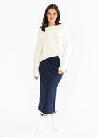 A woman modeling a cream off-the-shoulder knit sweater and a dark navy midi skirt, paired with white ankle boots, against a plain white studio background.