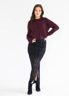 A woman modeling a dark burgundy knit sweater and a black and gray plaid skirt with sheer mesh panels and a front slit, paired with black ankle boots, against a plain white studio background.
