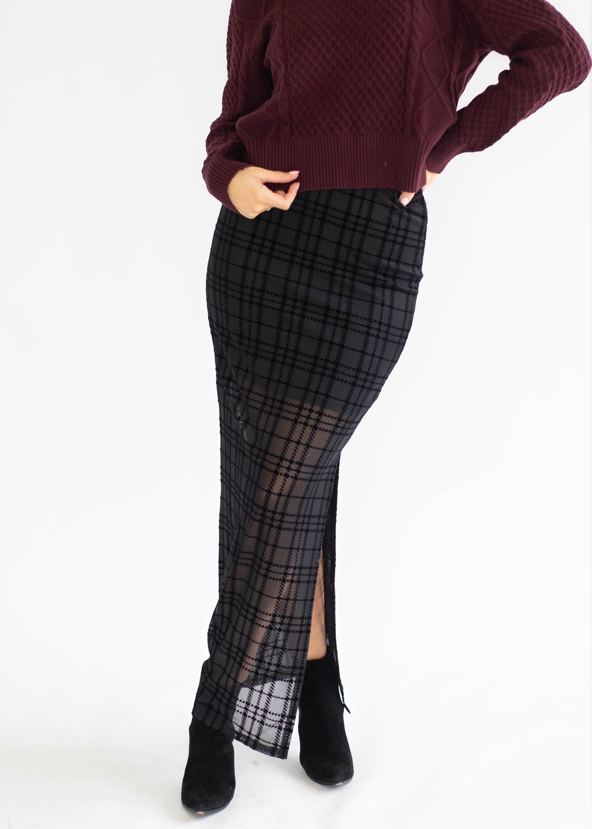 A model wearing a black and gray plaid midi skirt with a sheer mesh overlay and side slit, paired with a burgundy knit sweater and black ankle boots, against a plain white background.