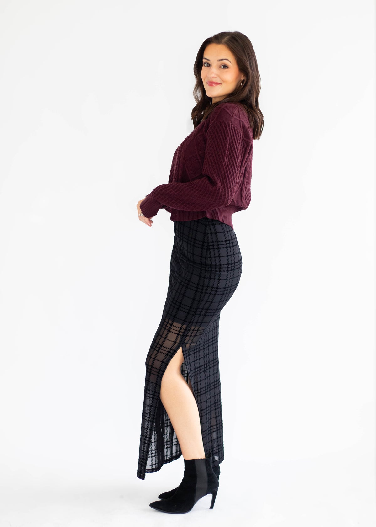A woman modeling a dark burgundy knit sweater and a black plaid midi skirt with a sheer thigh-high slit, paired with black ankle boots, against a plain white studio background.