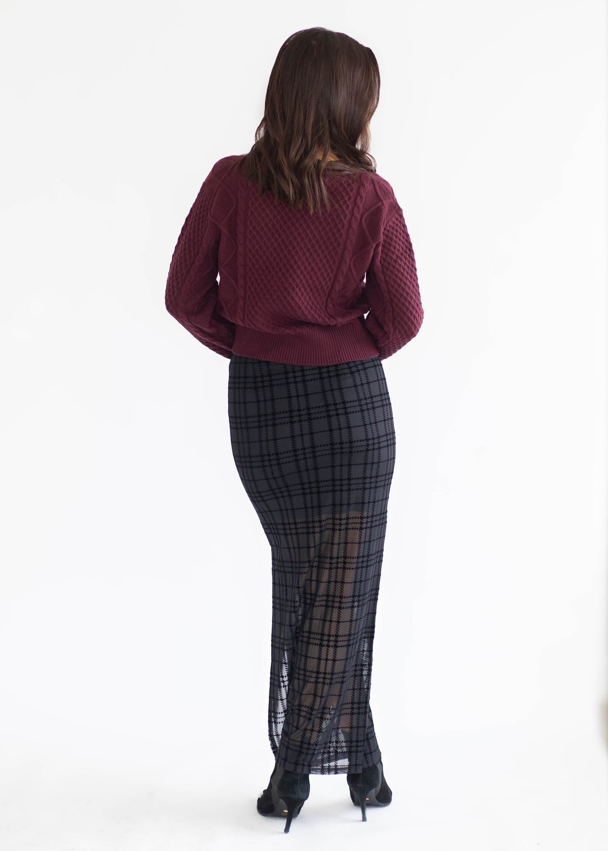 A woman viewed from behind wearing a burgundy knit sweater and a black and gray plaid maxi skirt with sheer tulle panels, set against a plain white studio background.