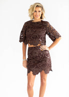 A woman modeling a dark brown lace two-piece set consisting of a short-sleeve crop top and a matching mini skirt, photographed against a plain white studio background.