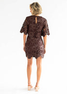 A woman viewed from the back wearing a short lace dress in a dark brown color, paired with beige block-heel sandals, against a plain white studio background.