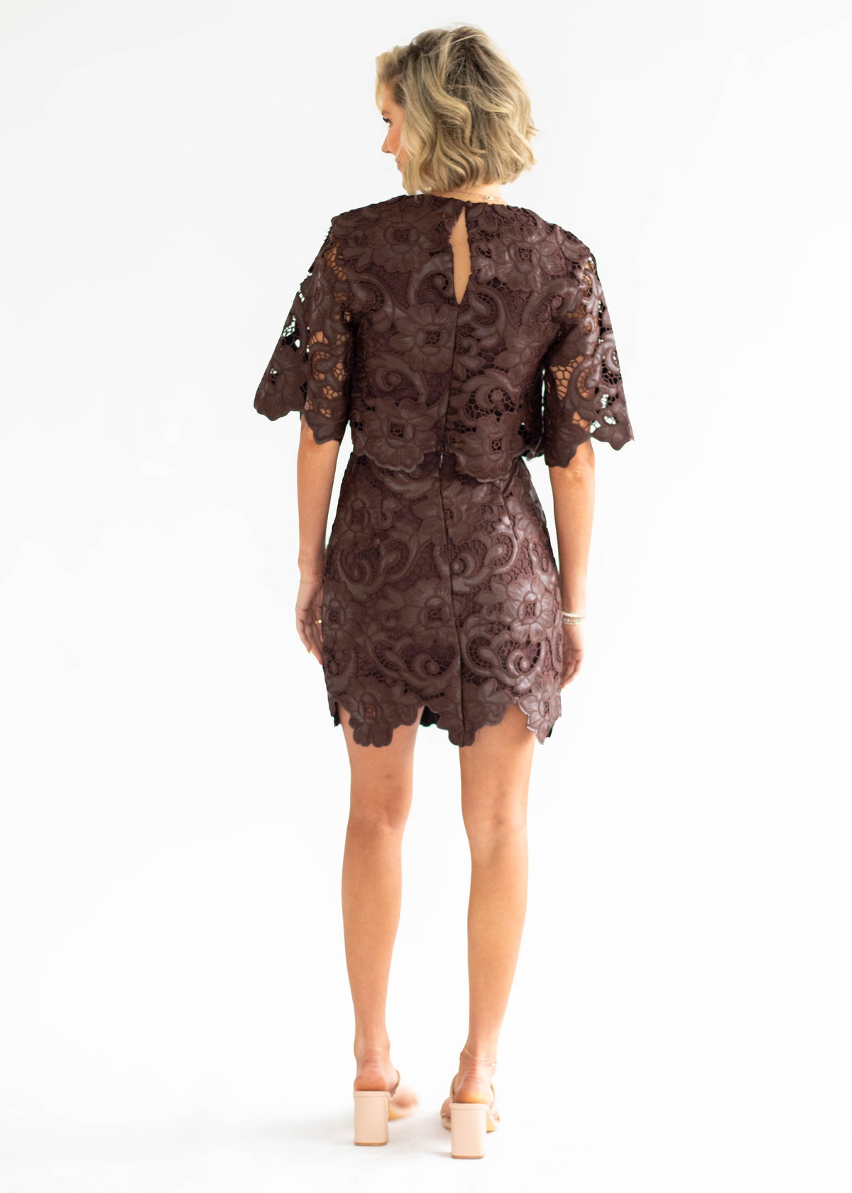 A woman viewed from the back wearing a short lace dress in a dark brown color, paired with beige block-heel sandals, against a plain white studio background.