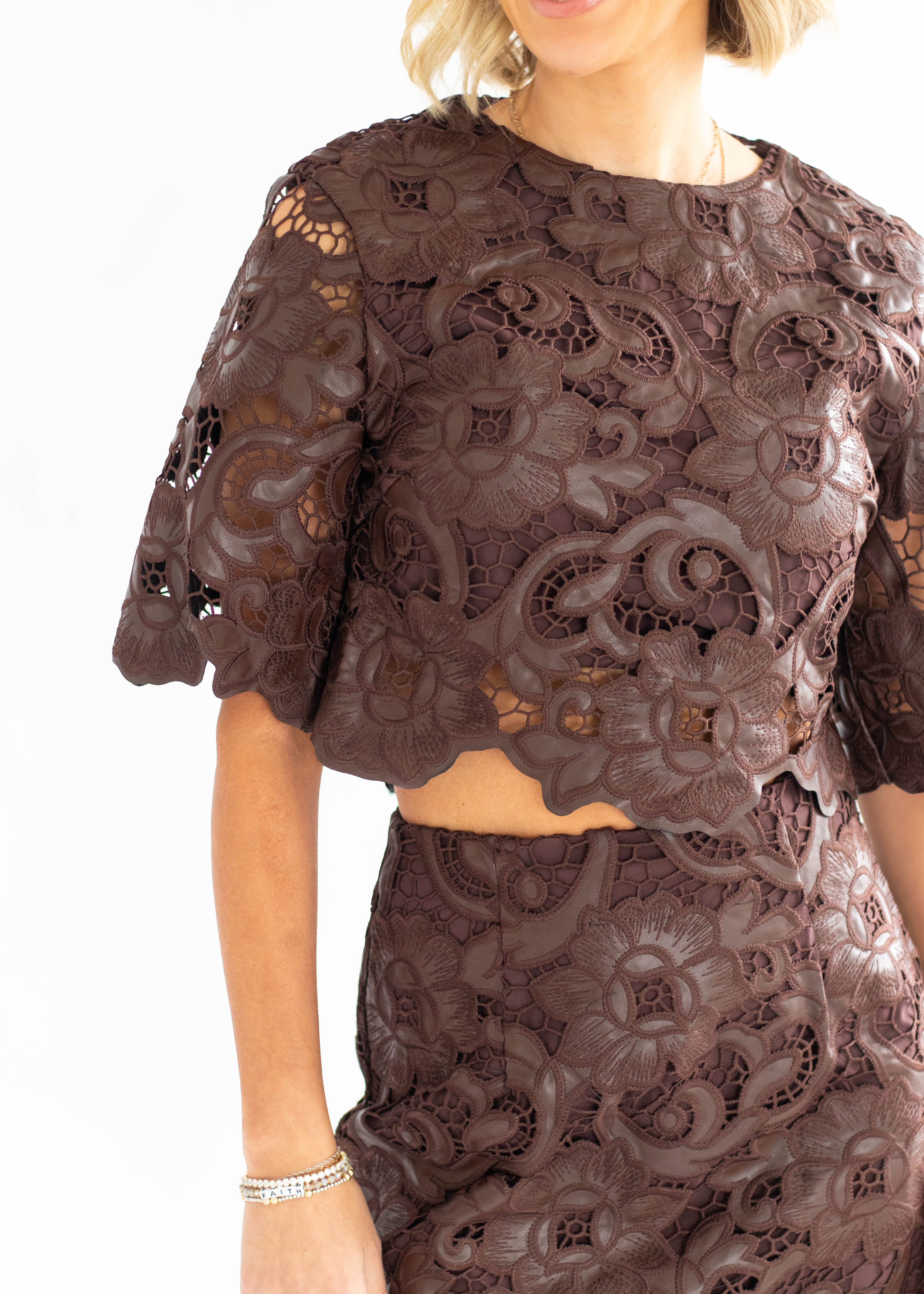A close-up of a model wearing a dark brown lace two-piece set, featuring a cropped short-sleeve top and a matching skirt, against a plain white background.