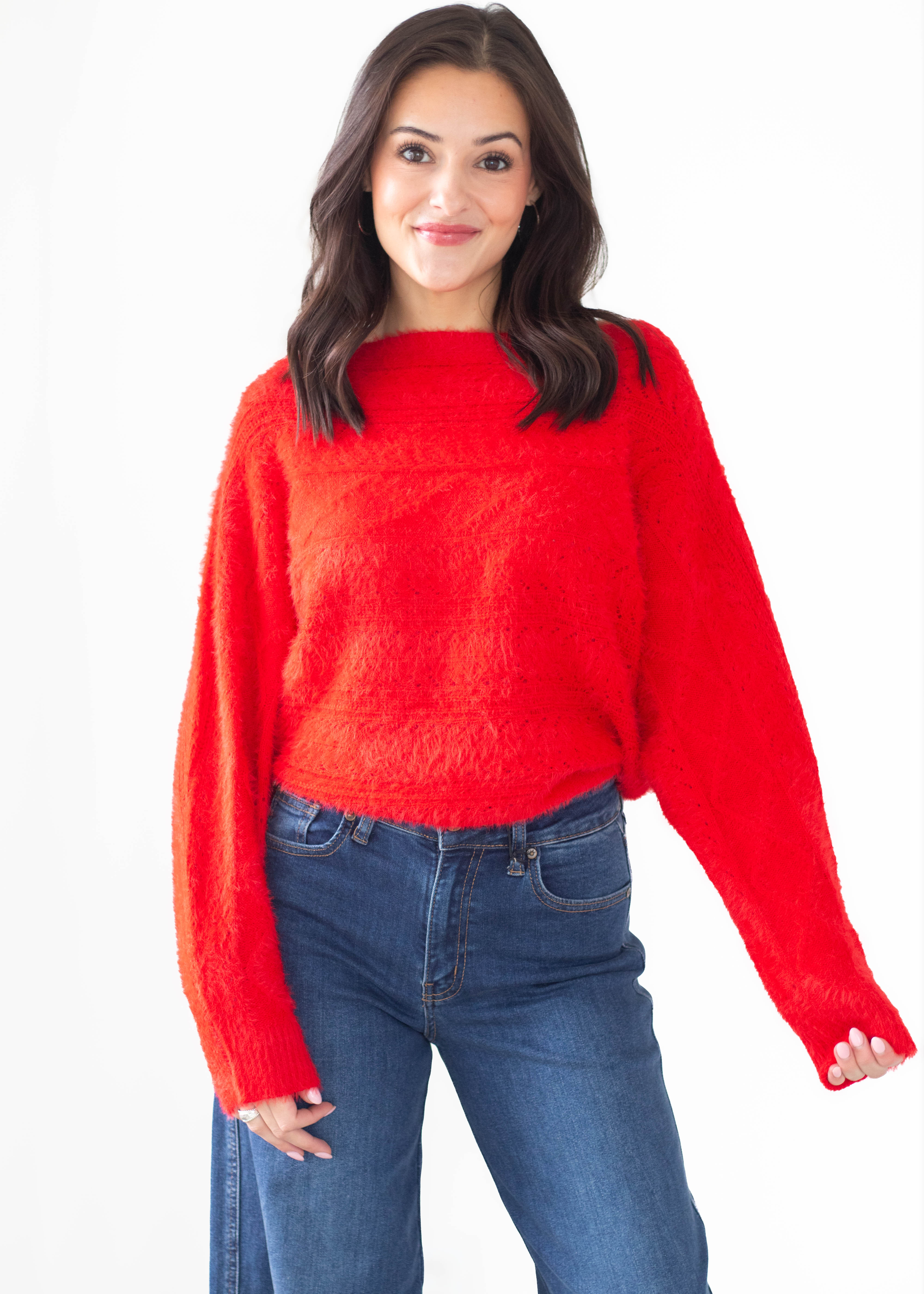 A woman modeling a bright red fuzzy knit sweater with a wide boat neckline and long sleeves, paired with blue jeans, against a plain white background.