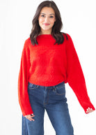 A woman modeling a bright red fuzzy knit sweater with a wide boat neckline and long sleeves, paired with blue jeans, against a plain white background.