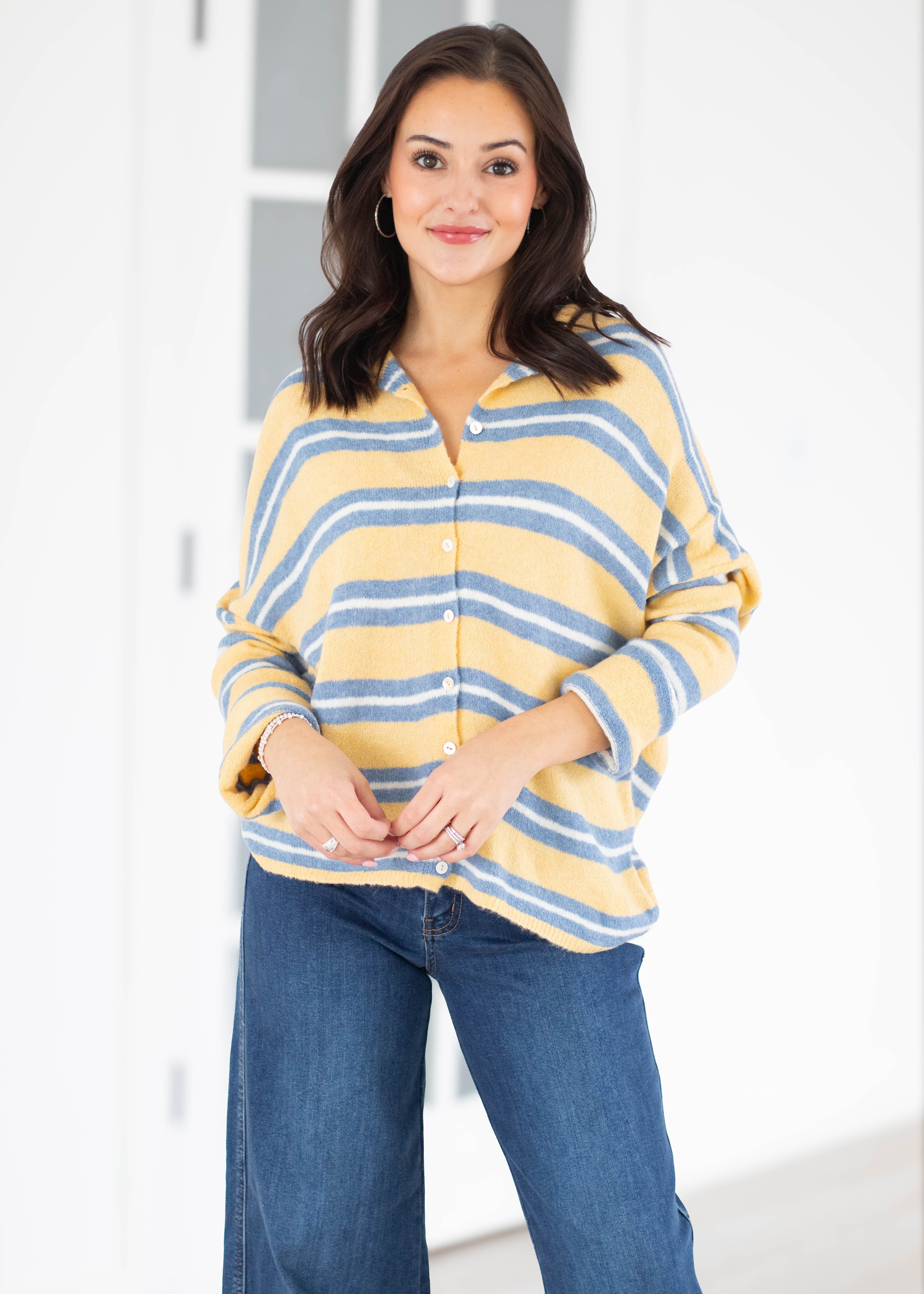 A woman modeling a yellow and blue striped button-up knit top with blue jeans, standing in a bright indoor setting.