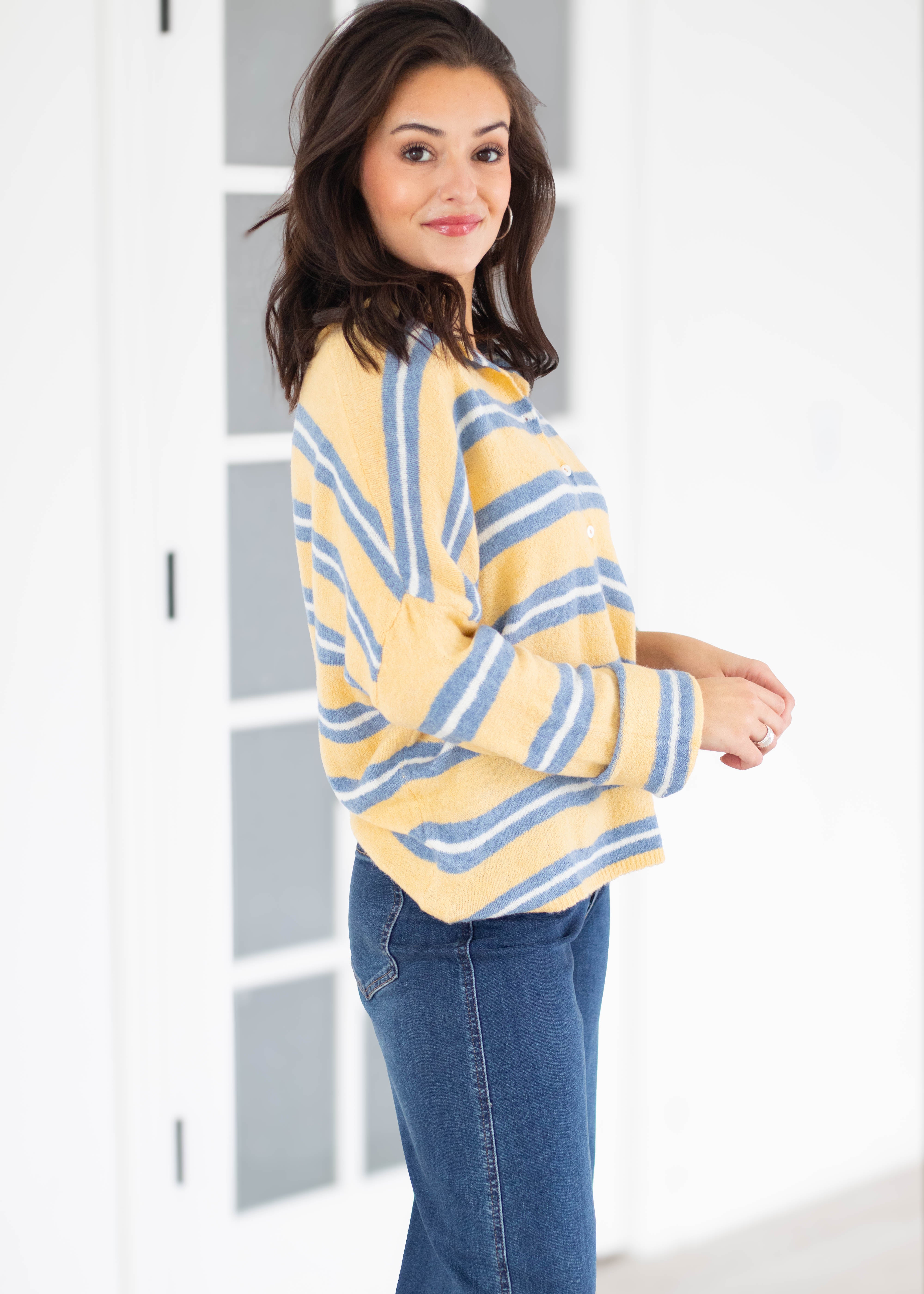 A woman wearing a yellow and blue striped knit cardigan sweater and blue jeans, standing in a bright white room with a glass-paneled door.