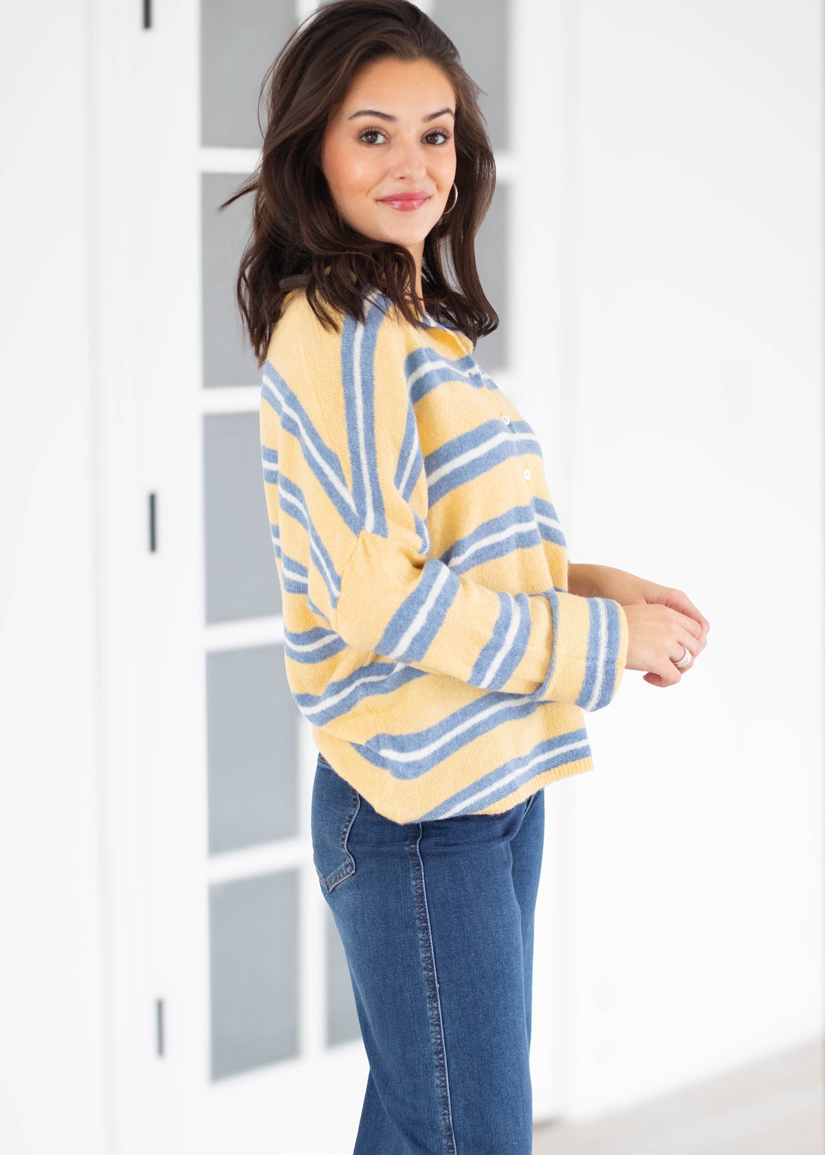 A woman wearing a yellow and blue striped knit cardigan sweater and blue jeans, standing in a bright white room with a glass-paneled door.