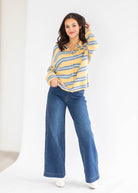 A woman modeling a yellow and blue striped long-sleeve button-up shirt with blue wide-leg jeans, standing against a clean white studio background.