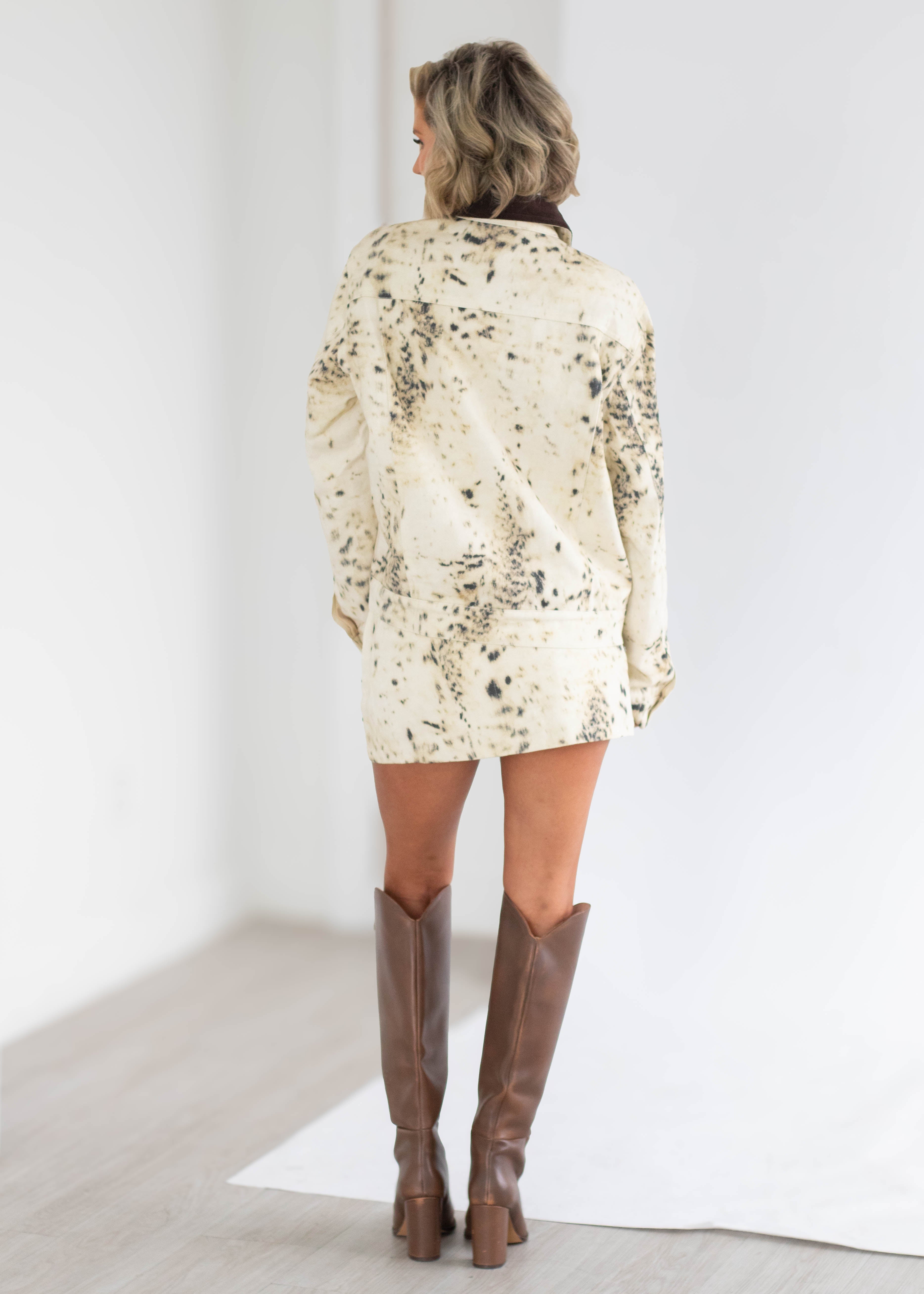 A woman viewed from behind wearing a cream-colored long-sleeve shirt with a black abstract paint splatter pattern and brown leather knee-high boots, standing in a bright white studio setting.