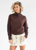 A woman wearing a dark brown ribbed turtleneck sweater and a white tie-dye mini skirt, standing against a plain white studio background.