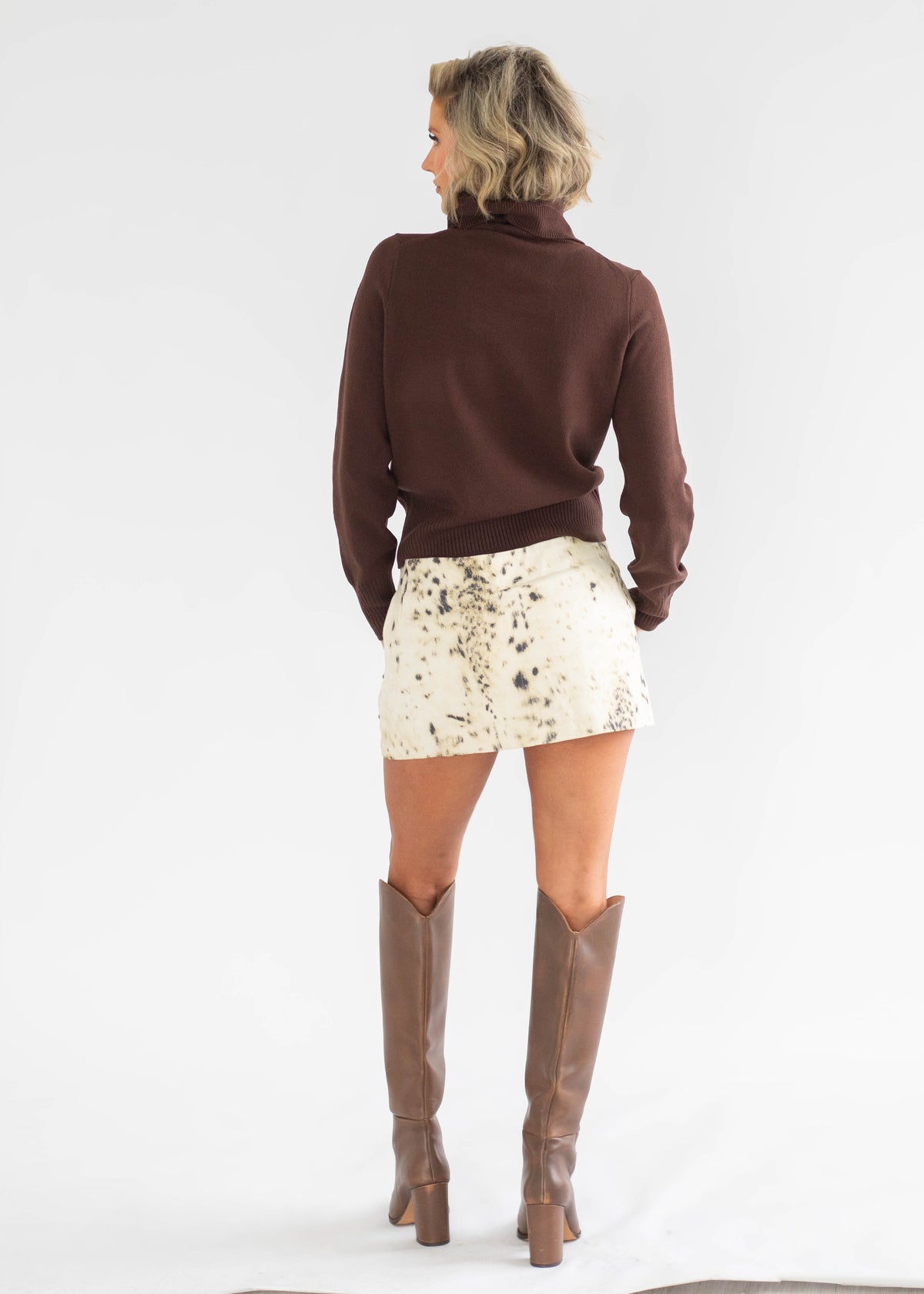A woman shown from the back wearing a dark brown turtleneck sweater, a cream snakeskin-patterned mini skirt, and brown knee-high boots, photographed against a seamless white studio background.