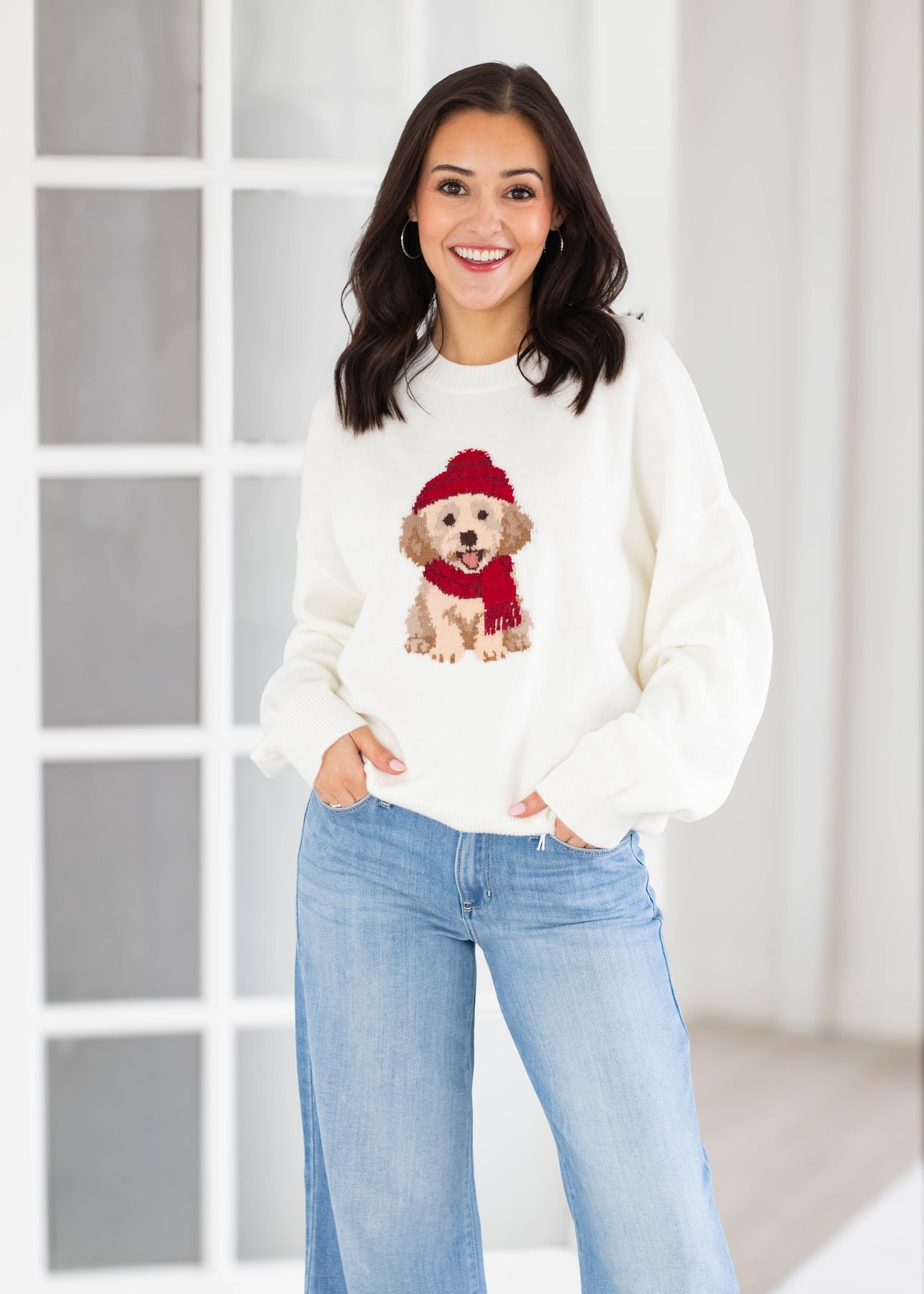 A woman wearing a cream-colored crewneck sweater with a graphic of a golden retriever dog wearing a red knit hat and red knit scarf, paired with light blue jeans.