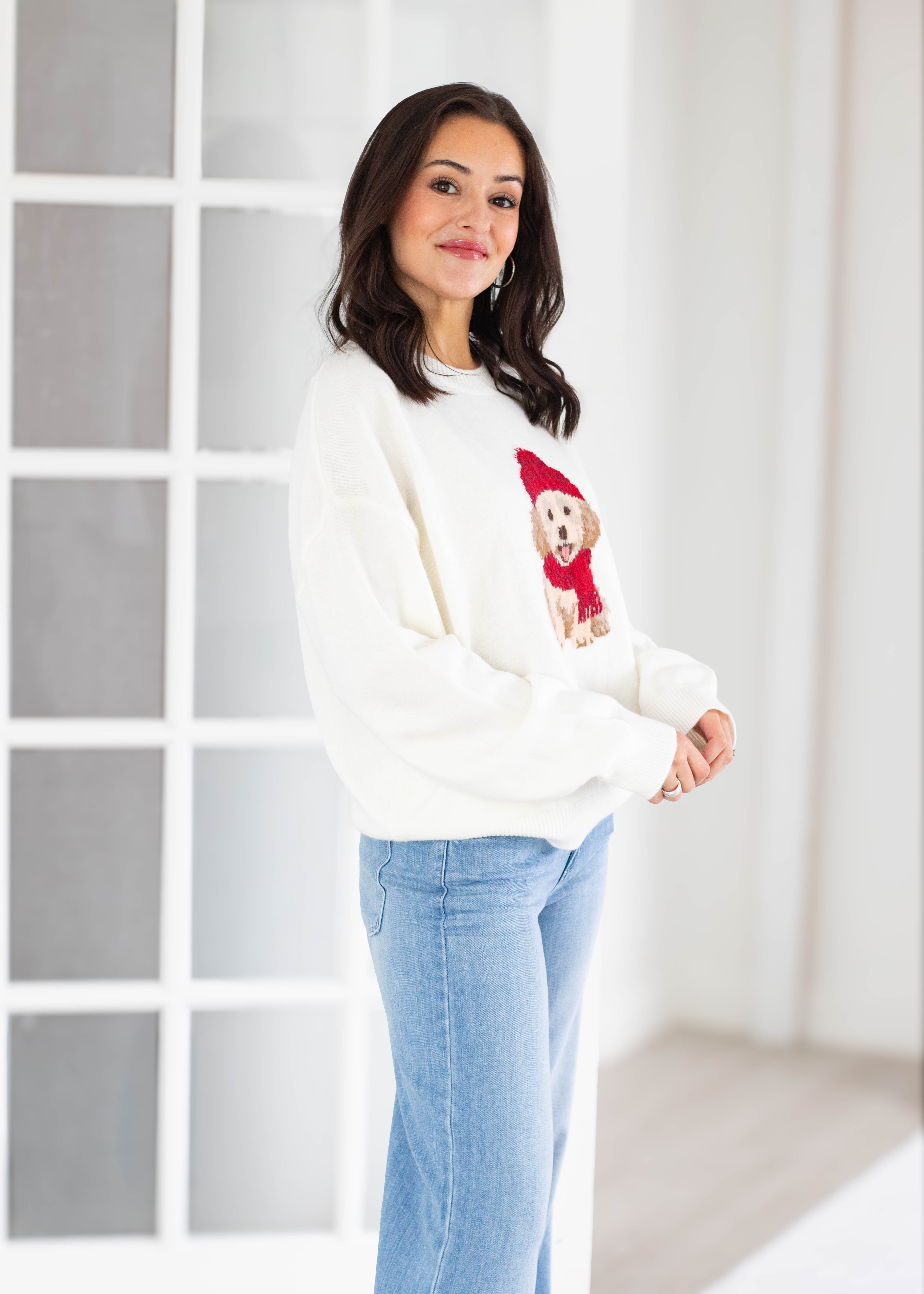 A woman wearing a white crewneck sweatshirt with a dog graphic and light blue jeans, standing in a bright, modern room with white paneled walls.