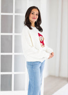 A woman wearing a white crewneck sweatshirt with a dog graphic and light blue jeans, standing in a bright, modern room with white paneled walls.