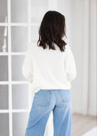 A woman viewed from behind wearing a cream-colored knit sweater and light blue wide-leg jeans, standing in front of a white paneled window.