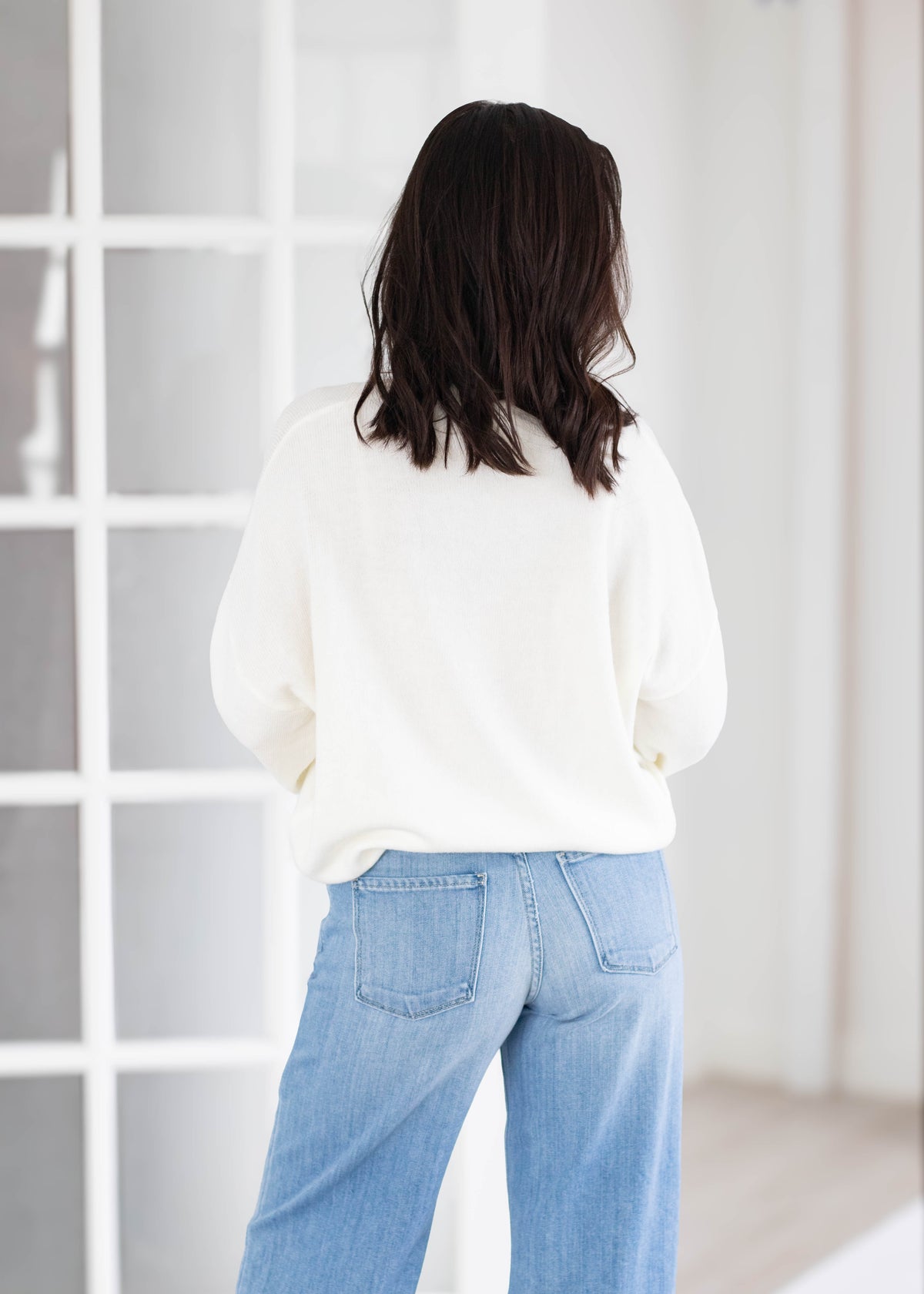 A woman viewed from behind wearing a cream-colored knit sweater and light blue wide-leg jeans, standing in front of a white paneled window.