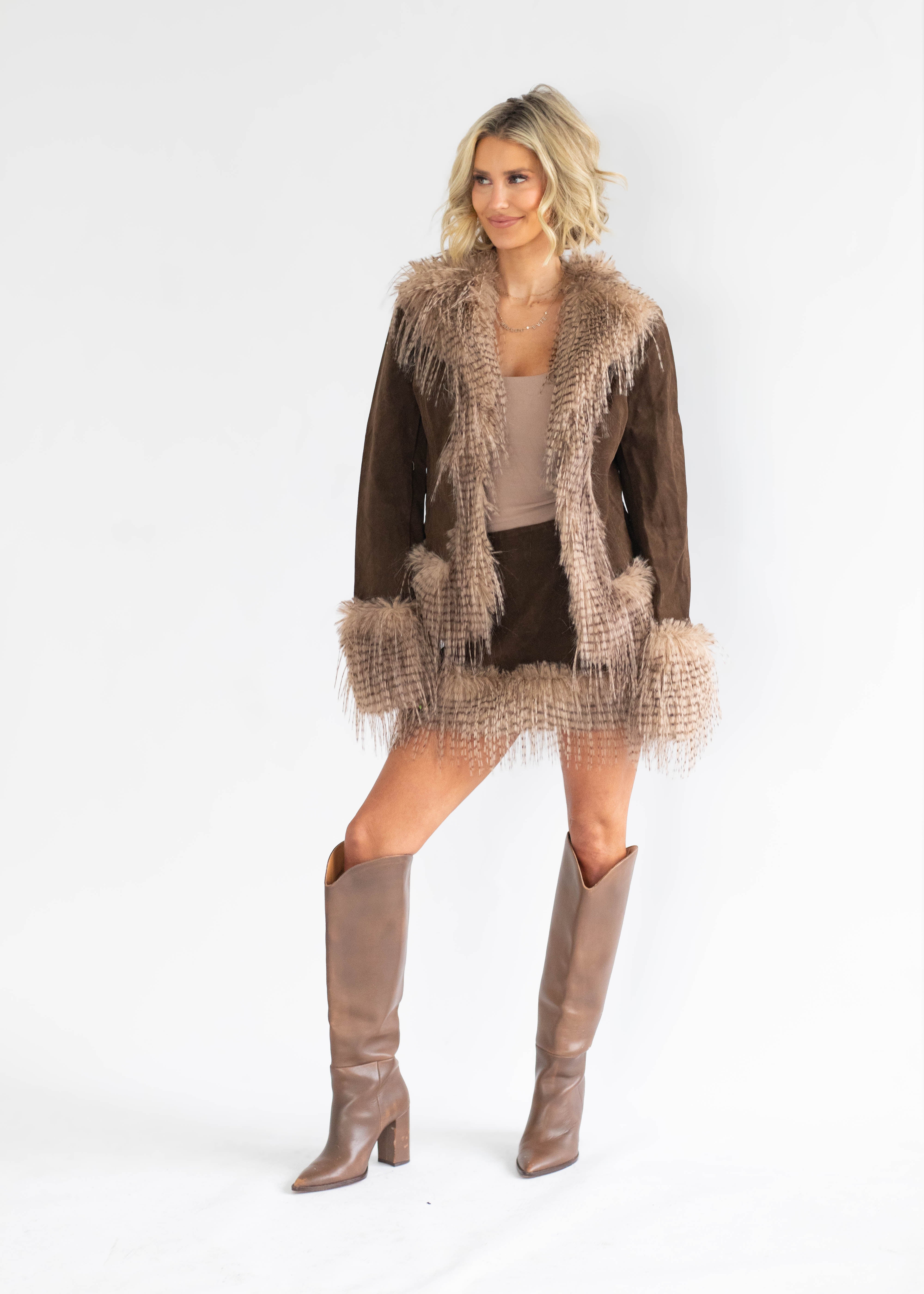 A woman modeling a brown suede jacket with beige faux fur trim, paired with a beige top, black skirt, and brown knee-high boots, against a plain white studio background.