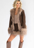 A woman modeling a brown suede jacket with beige faux fur trim, styled with a beige top, matching shorts, and beige cowboy boots against a plain white background.