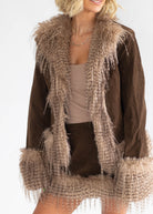 A woman wearing a brown suede jacket with a large beige faux-fur collar, cuffs, and hem, styled over a beige top against a plain white background.