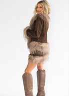 A woman modeling a brown suede jacket with beige fringe trim, paired with brown knee-high boots, against a plain white studio background.