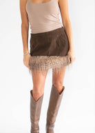 A model wearing a brown mini skirt with a fringe hem, paired with a beige tank top and brown knee-high boots, shown from the chest down against a white background.