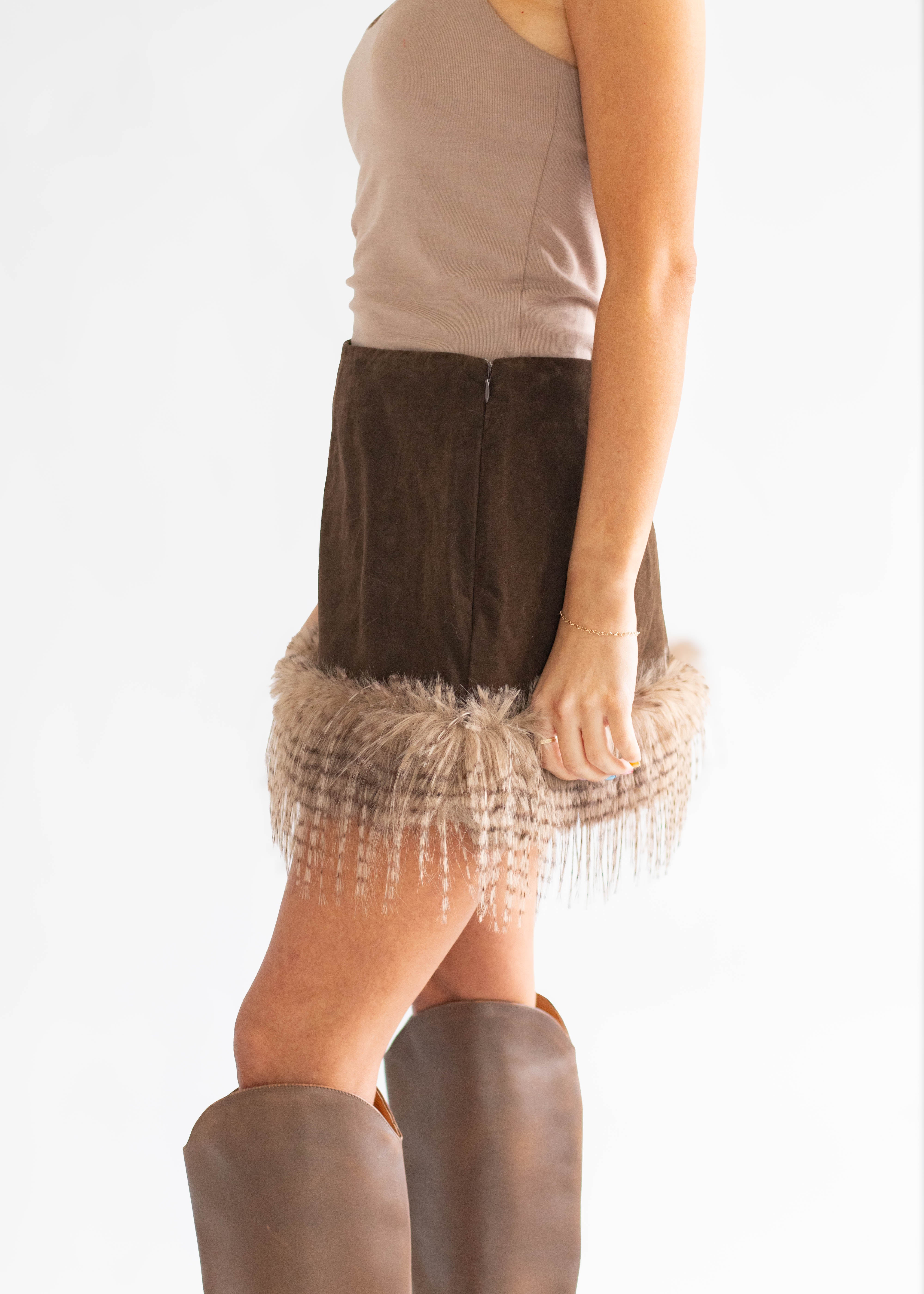 A model shown from the side wearing a dark brown suede mini skirt with a fringed hem, paired with a beige tank top and brown knee-high boots, against a plain white background.
