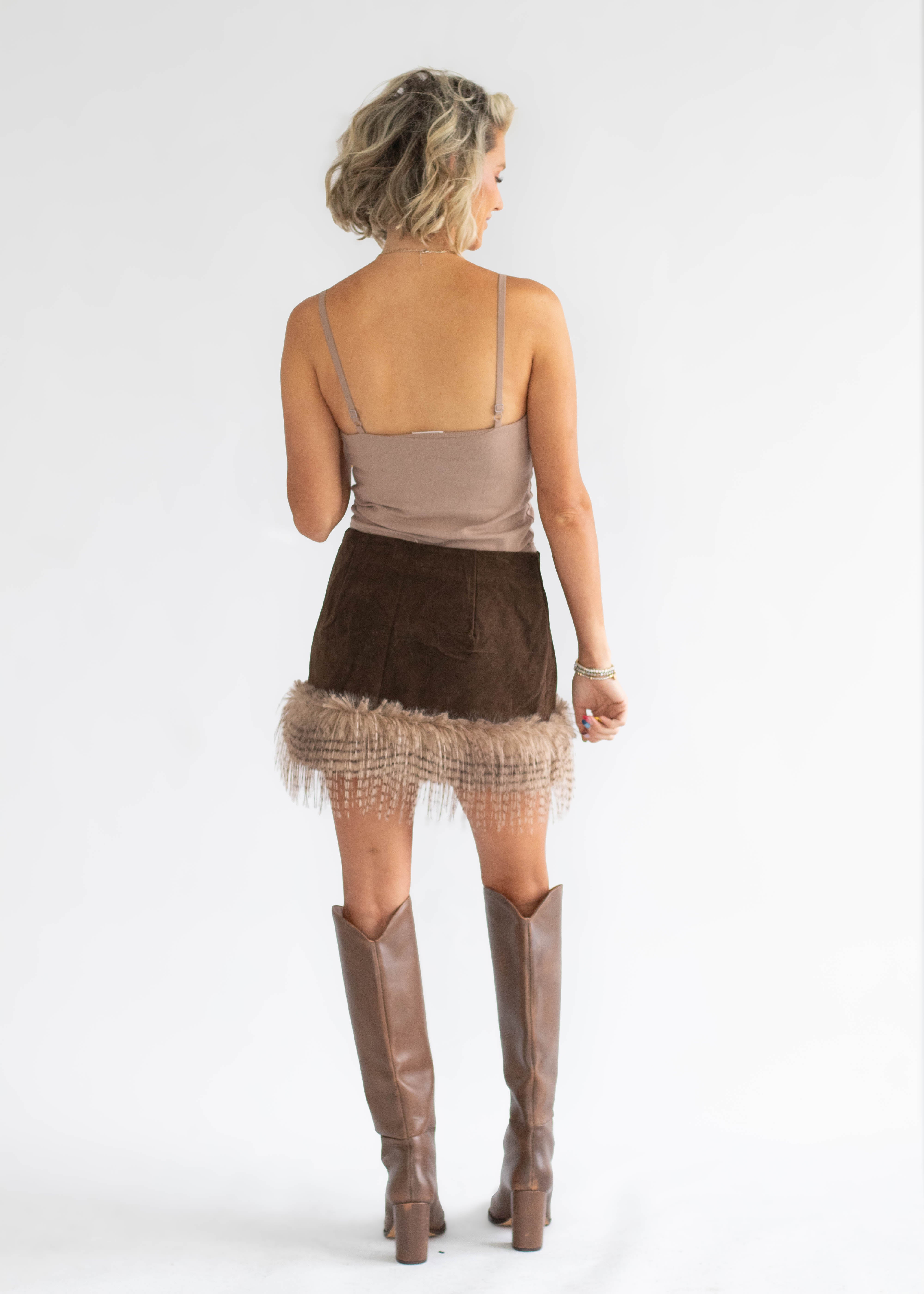 A woman shown from the back wearing a taupe spaghetti-strap top, a brown mini skirt with a fringed hem, and brown knee-high boots, photographed against a light gray studio background.