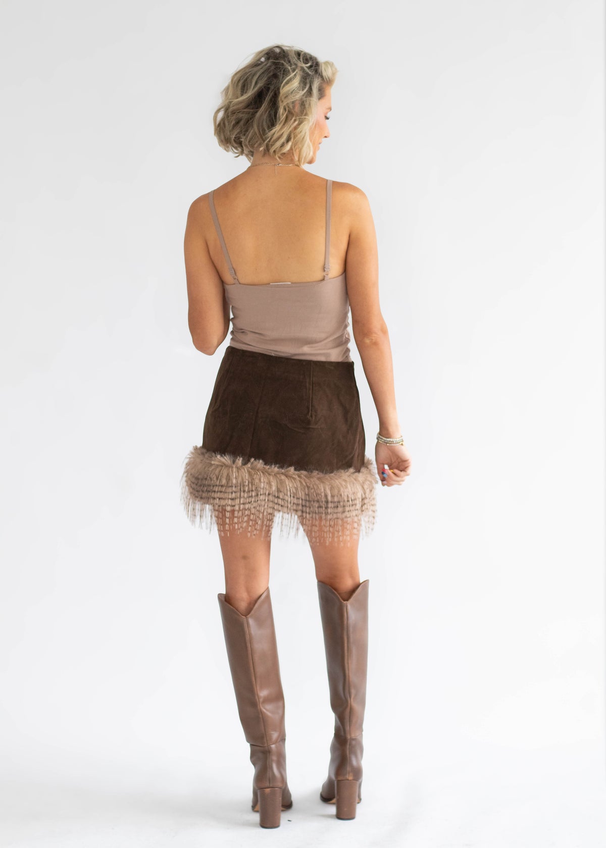 A woman shown from the back wearing a taupe spaghetti-strap top, a brown mini skirt with a fringed hem, and brown knee-high boots, photographed against a light gray studio background.