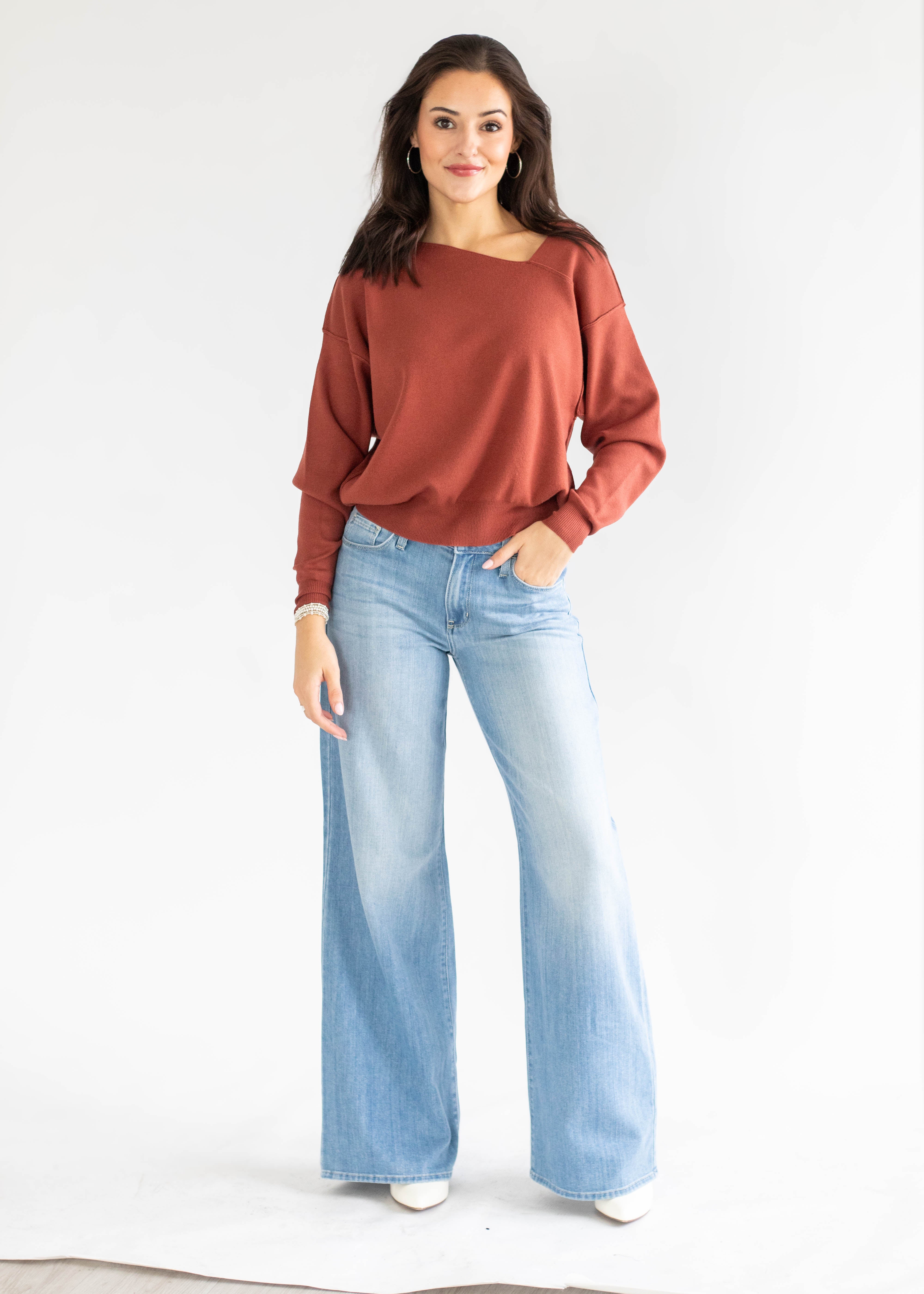 A woman modeling a rust-colored, off-the-shoulder long-sleeve top paired with light-wash wide-leg jeans and white shoes, standing against a plain white studio background.